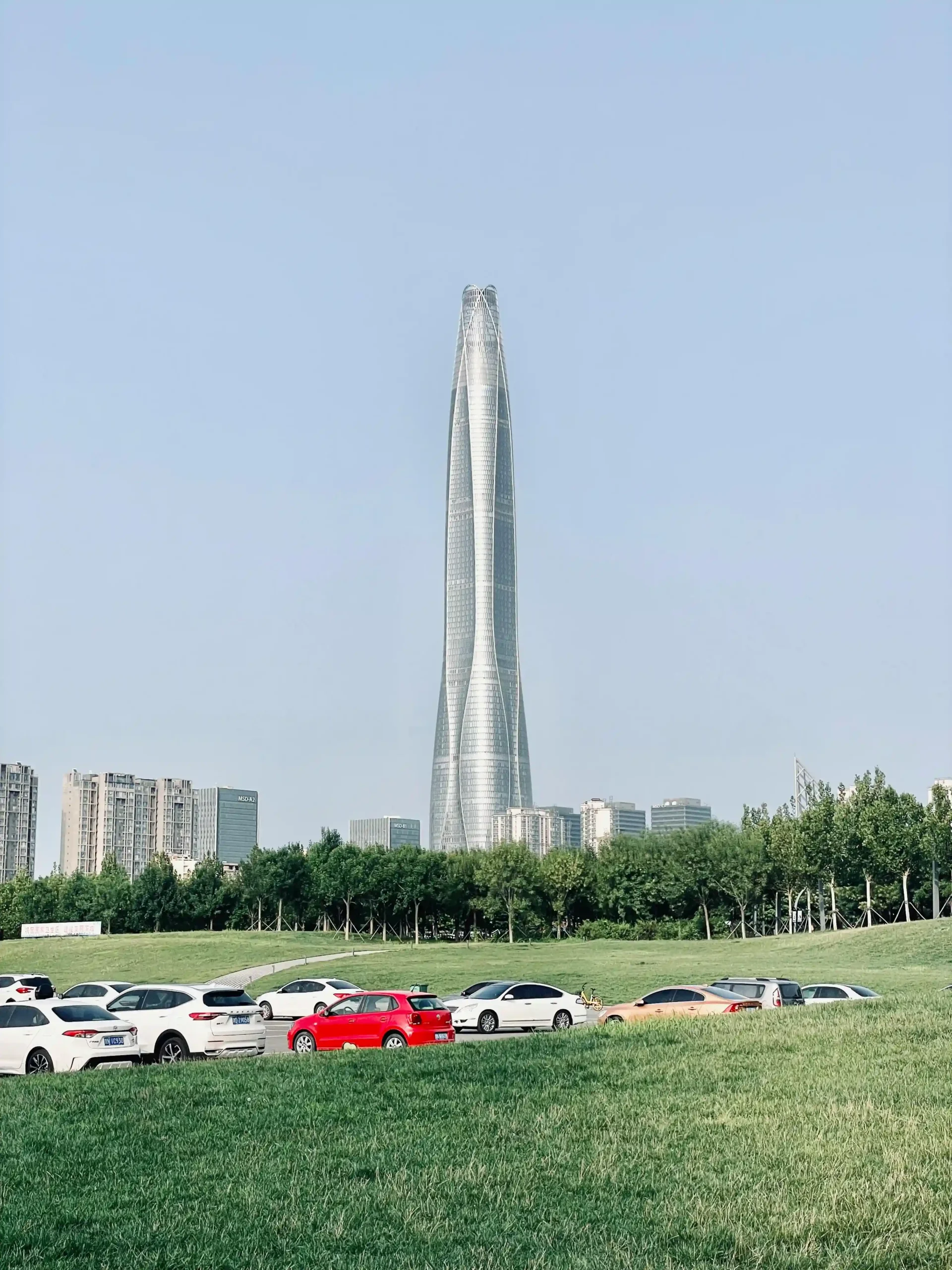 10 Tallest Buildings in the World (2025) Tianjin CTF Finance Centre in the distance, surrounded by green grass and parked cars, under a clear blue sky.